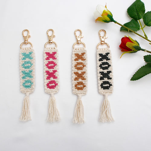 Four white woven keychains with woven text patterns and tassels, in blue, pink, tan, and black colors, and gold clasps. Displayed alongside a red and white rose, on a white background.