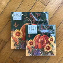 Load image into Gallery viewer, Matching cocktail and dinner sized paper napkin sets wrapped in plastic packaging with a white label in the upper left corner. White label has &quot;ihr&quot; and &quot;SINCE 1977&quot; in black print. Napkins have a harvest theme including an orange pumpkin, yellow sunflowers, a green and purple cabbage, red berries, and an orange butterfly,  on a tan and black background. Napkins are lying on a brown wooden plank surface.