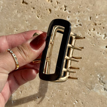 Load image into Gallery viewer, Black rectangular metal claw clip with gold hardware held in a hand against a beige travertine surface with soft shadows. 