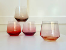 Load image into Gallery viewer, Set of four hand-blown crystal stemless glasses in gradient shades of red, plum, amber, and rose, arranged on a white surface against a white background, with one glass stacked on top of another. 