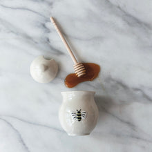 Load image into Gallery viewer, White honey jar with a bee design , its lid, a wooden spoon , and spilled honey on a marble surface.