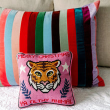 Load image into Gallery viewer, Pink needlepoint pillow featuring an orange tiger face with yellow eyes and a pink nose, white stars, and navy botanical motifs, with &quot;Merry Christmas Ya Filthy Animal&quot; stitched in red, displayed in front of a large multi-colored striped pillow on a neutral fabric sofa.