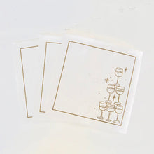 Load image into Gallery viewer, Set of three white cotton cocktail napkins with a gold border, featuring a gold outlined stack-of-glasses illustration on the top napkin, displayed against a white background. 