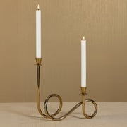 Two-tier brass candle holder in a polished gold finish with a looping base, holding two white lit taper candles. Displayed against a beige-colored background.