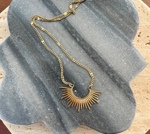 Gold necklace featuring a bold sunburst pendant on a delicate chain, displayed on a quatrefoil-shaped gray stone tile layered over a beige travertine slab on wooden surface.