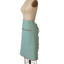 Load image into Gallery viewer, Holiday Apron, green cotton gingham waist apron displayed on a light tan dress form, with waist ties tied at the back and red scalloped trim along the hem and front pocket and partially visible at the back, against a white background. 