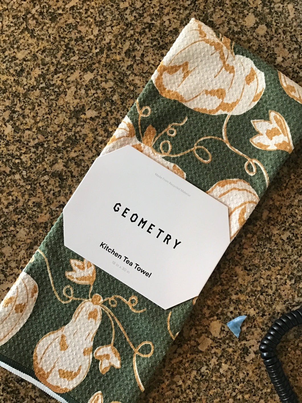 A green kitchen tea towel with a pumpkin trail design, featuring white and orange gourds and leaves pattern, laid out flat with a label that reads 'GEOMETRY'.