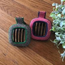 Load image into Gallery viewer, Two beaded hair clips laid on a wooden surface, one clip with a green ,black, and white color beads and the other with a red, blue, and pink color beads. Green foliage on the right side.