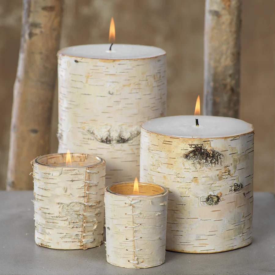 Assorted lit birch-wrapped candles displayed on a gray surface against a neutral background with birch branches. 