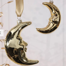Load image into Gallery viewer, Gold Crescent Moon Ornaments, lifestyle image of two reflective gold-tone blown glass crescent moons with sculpted facial profiles facing each other, with the larger moon hanging from a wide gold ribbon beside delicate gold decorations against a pale background.