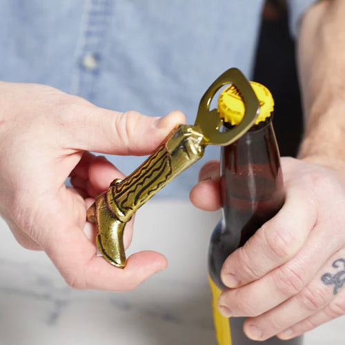 Man's hands opening a beer bottle with a gold bottle opener in the shape of a cowboy boot. Background is the mans chest in a light blue shirt over a white marble countertop. 