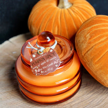 Load image into Gallery viewer, Candle in a pumpkin colored glass with a tag visible, Glass is ribbed shaped. Tag is red with the text: &quot;PADDYWAX&quot;, &quot;PUMPKIN SPICE&quot;, &quot;notes of apple &amp;vanilla&quot; , and &quot;SOY WAX CANDLE&quot;. Candle is on a wooden surface with two pumpkins beside it . Against a black background.