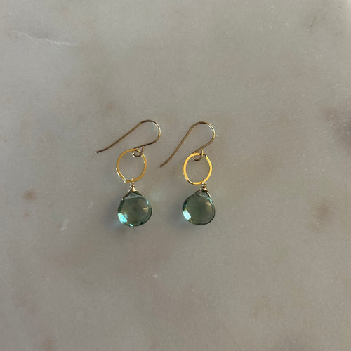 A pair of gold drop earrings with a teardrop aquamarine gem on a gold ring. Marble white background. 