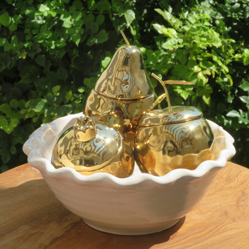 Gilded ceramic fruit candles in lidded jar shapes of pear, citrus, and pomegranate, displayed together in a white scalloped bowl on a wooden surface with green foliage in the background. 