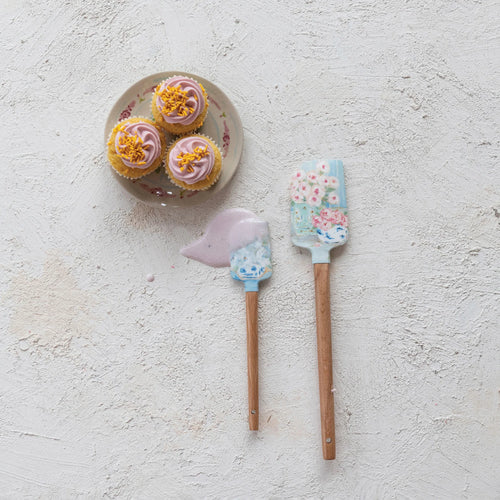 A big and small baby blue silicone spatula, with floral designs on the silicone end and wooden handles. Displayed alongside three pink frosted cupcakes on a small cream colored plate with a red flower pattern.  Small spatula has a pale pink cream on it and on the white textured surface.  
