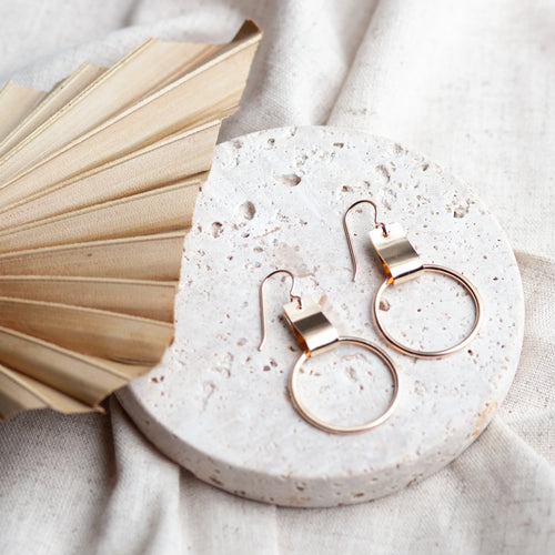Pair of rose gold earrings with a drop hoop on a porous white pedestal on a white linen sheet. To the side of the pedestal is a tan fan. 