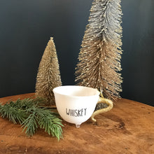 Load image into Gallery viewer, A white teacup ornament with a gold glitter handle, a clear footed bottom,  and &quot;WHISKEY&quot; in black text. A green fir tree cutting and two decorative frosted Christmas trees next to the teacup. On a wooden surface . Against a dark background. 