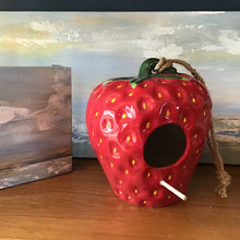 Load image into Gallery viewer, A red ceramic strawberry birdhouse with a tied rope for hanging and a wooden perch. Two ocean and sky paintings in the background, and on a wooden surface. 