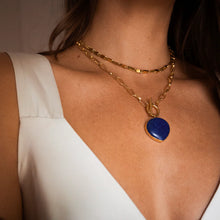 Load image into Gallery viewer, Model wearing a gold-plated brass chain necklace featuring a large blue lapis stone pendant with a toggle closure layered with a shorter gold chain, shown against a deep V-neck white sleeveless top.  