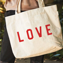 Load image into Gallery viewer, Natural cotton canvas tote bag with bold red "LOVE" lettering and shoulder straps, shown worn over the shoulder.