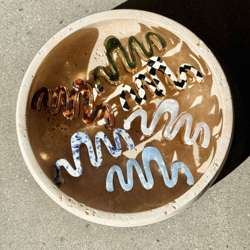 Six wavy claw clips in tortoiseshell, forest green, checkered black and white, pearl, blue speckle, and coastal blue displayed inside a round beige bowl on a textured surface with soft shadows.  