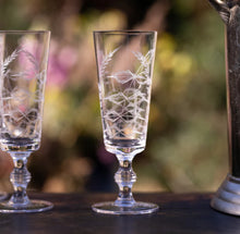 Load image into Gallery viewer, Two champagne flutes with leaf etchings on a wooden platform. Blurred nature background. 