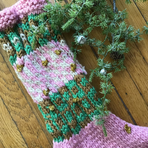 Crocheted Christmas stocking in pink, green, brown, and white yarn decorated with small gold glittery accents. Displayed on a wooden surface with green garland and silver jingle bells.  