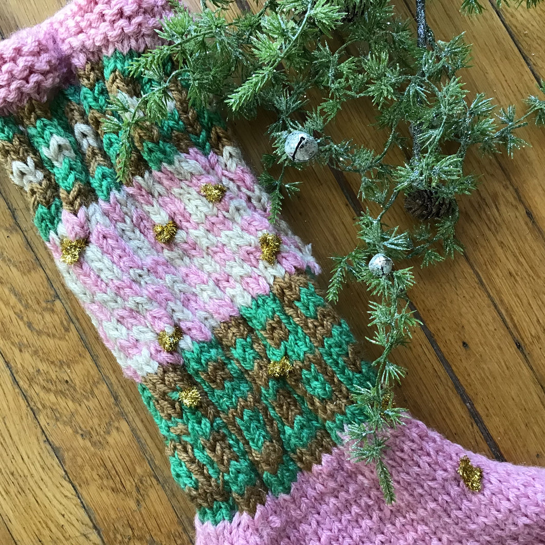 Crocheted Christmas stocking in pink, green, brown, and white yarn decorated with small gold glittery accents. Displayed on a wooden surface with green garland and silver jingle bells.  