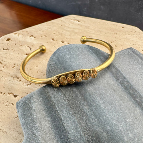 Gold-finish open cuff bracelet featuring a row of etched floral medallions, displayed on a gray and beige stone tile on a wooden surface. 