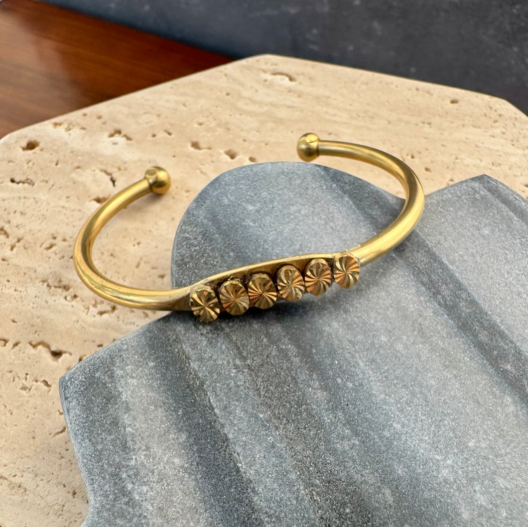 Gold-finish open cuff bracelet featuring a row of etched floral medallions, displayed on a gray and beige stone tile on a wooden surface. 