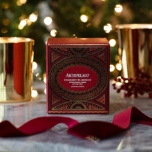 Load image into Gallery viewer, Garnet red and metallic gold Cranberry St. Germain candle box on a white marble surface with a red ribbon, metallic gold candle holders, and a lit blurred Christmas tree in the background.