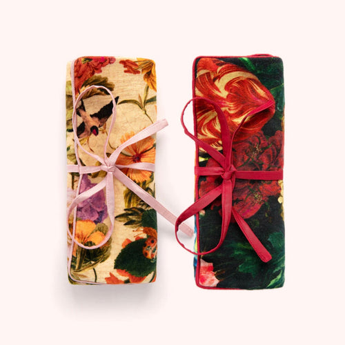 Two velvet jewelry rolls displayed side by side on a white background- one in a floral and bird pattern with a white base tied with a pink ribbon, and the other in a red and green floral pattern tied with a red ribbon. 