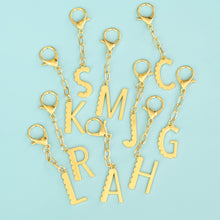 Load image into Gallery viewer, Set of gold-plated enamel initial bag charms featuring letters S, M, J, C, K, L, R, A, and H, each with a scalloped edge design, key ring, and a clasp, arranged in a scattered layout on a blue background. 