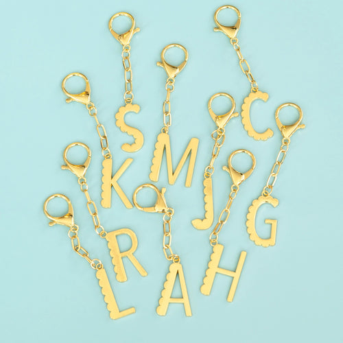 Set of gold-plated enamel initial bag charms featuring letters S, M, J, C, K, L, R, A, and H, each with a scalloped edge design, key ring, and a clasp, arranged in a scattered layout on a blue background. 