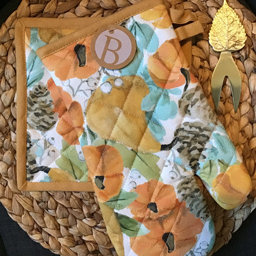Matching quilted potholder and oven mitt set on a brown woven surface with a gold appetizer fork with a leaf handle. 
Matching set is white with an orange pumpkin ,gourd, brown pinecone, white berry, and green leaf design , and an orange border and trim. Oven mitt has a brown round label with 