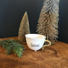 Load image into Gallery viewer, White teacup ornament with a gold glitter handle, a clear footed bottom, and  &quot;Wine&quot; in black text. A green fir tree cutting and two decorative frosted Christmas trees next to the teacup. On a wooden surface. Against a dark background. 