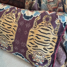 Load image into Gallery viewer, Tiger Lumbar Pillow, close-up angled view of plum cotton velvet blend lumbar pillow with orange tiger motif, teal, blue, and beige geometric accents, and burgundy fringe trim on light-colored fabric surface with coordinating fabric in background. 