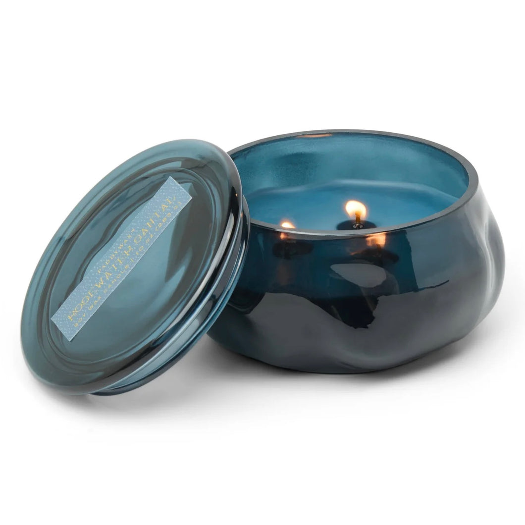 Blue lit candle in a blue glass container. Blue candle lid propped against candle. Against a white background.