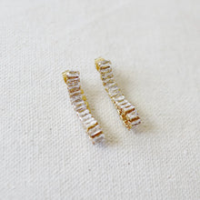 Load image into Gallery viewer, Pair of gold IP-plated earrings with curved rows of clear baguette-cut CZ stones displayed on a soft white textured background.