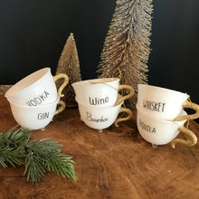 Load image into Gallery viewer, Collection of six white holiday teacup ornaments with gold glitter handles and a clear footed bottom.  Each teacup has a word in black text- &quot;VODKA&quot;, &quot;GIN&quot;, &quot;Wine&quot;, &quot;Bourbon&quot;, &quot;WHISKEY&quot;, and  &quot;TEQUILA&quot;.  Teacups stacked in twos on a wooden surface. A tall and mini decorative frosted Christmas tree behind the cups , and a green fir tree cutting in front .  Against a dark background.   