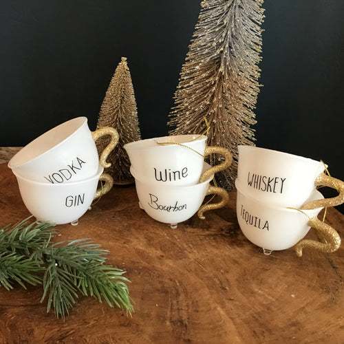 Collection of six white holiday teacup ornaments with gold glitter handles and a clear footed bottom.  Each teacup has a word in black text- 