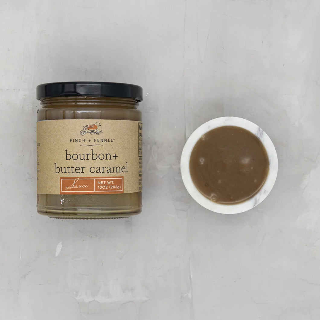 A jar of bourbon + butter caramel sauce next to a small bowl filled with the sauce, placed on a light-colored surface.