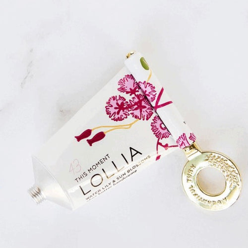 A white tube of lotion with a pink and red floral design and a gold key-shaped tool to squeeze all lotion out, on a white marble surface. Tube has brand name in black: 