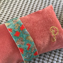 Load image into Gallery viewer, Pink cushion with an embroidered &quot;P&quot; , and a teal beaded band bracelet with red, brown, and green leopard spots wrapped around it ,on a gray textured surface.