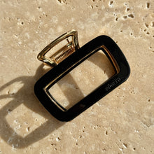 Load image into Gallery viewer, Black rectangular metal claw clip with gold hardware displayed on a beige travertine surface with soft shadows. 