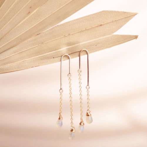 A pair of long earrings with two gold link chains and drop light blue gems. The earrings are in a tan fan on a white background. 