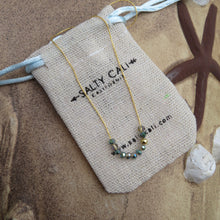 Load image into Gallery viewer, Gold necklace with iridescent  beads on a &#39;Salty Call&#39; branded pouch on sand with starfish