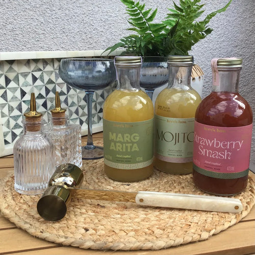 Bottles of Margarita, Mojito, and Strawberry Smash with a cocktail glasses, 2 glass bottles and jigger on a woven tray, tile and stucco backsplash. 