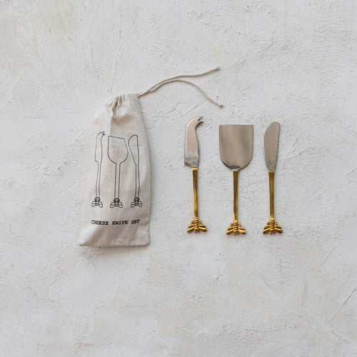 A set of three cheese knives with gold finish bee handles, accompanied by a white fabric storage bag with 3 cheese knives printed in black, displayed against a white textured  background.