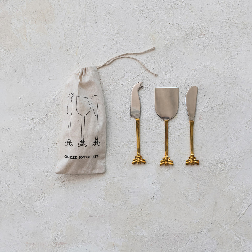 A set of three cheese knives with gold finish bee handles, accompanied by a white fabric storage bag with 3 cheese knives printed in black, displayed against a white textured  background.